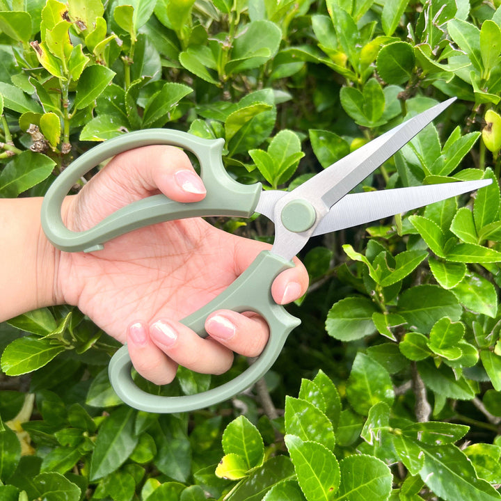 Hand holding green gardening shears against a green leafy bush by House of Agave.