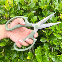 Hand holding green gardening shears against a green leafy bush by House of Agave.